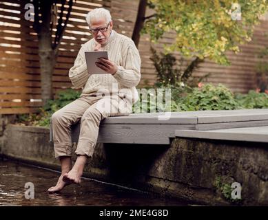 Senior man sitting by garden pond using digital tablet Stock Photo
