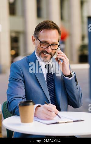 Smiling businessman talking over smart phone and writing on clipboard at cafe Stock Photo
