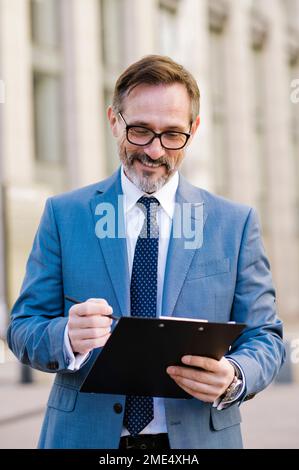 Smiling mature businessman standing with clipboard outside building Stock Photo