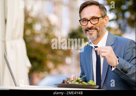 Smiling mature businessman eating salad with laptop on table at cafe Stock Photo