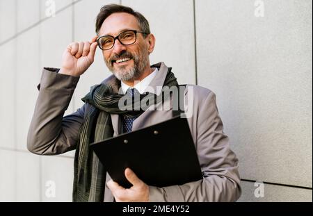 Happy mature businessman adjusting eyeglasses in front of wall Stock Photo
