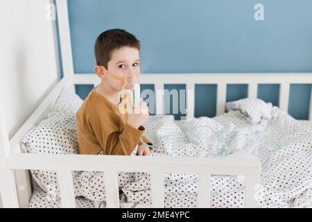 Boy wearing nebulizer mask sitting on bed at home Stock Photo