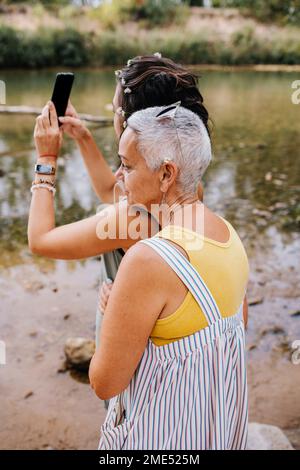 Women taking selfie through mobile at lakeshore Stock Photo