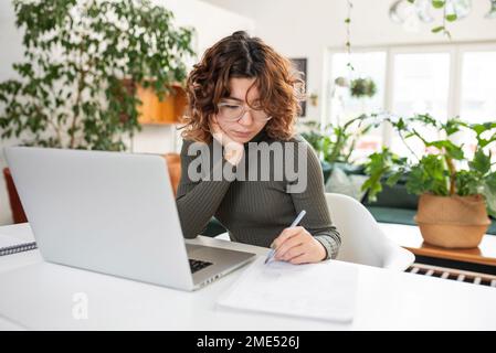 Freelancer writing notes sitting in front of laptop at home Stock Photo