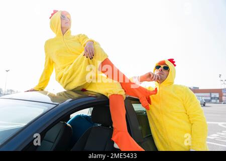 Woman in yellow chicken costume sitting on car with thoughtful man in parking lot Stock Photo