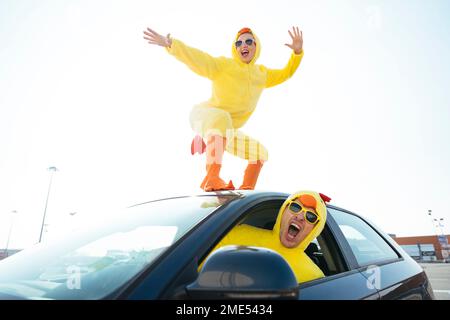 Man screaming and driving car with carefree woman in chicken costume dancing on roof Stock Photo