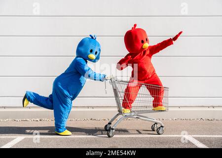 Friends wearing red and blue duck costumes having fun with shopping cart by wall Stock Photo