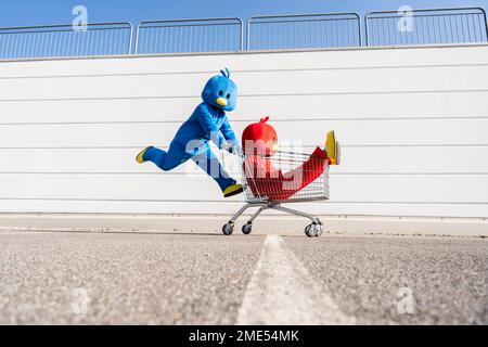 Man and woman wearing duck costumes playing with shopping cart by wall Stock Photo