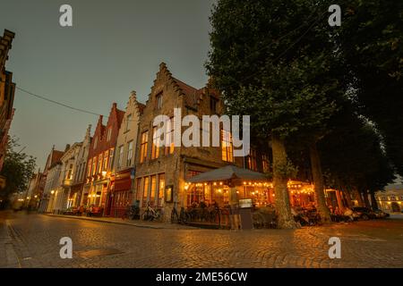 Belgium, West Flanders, Bruges, Cobblestone street stretching in front of sidewalk cafe and row of town houses at dusk Stock Photo