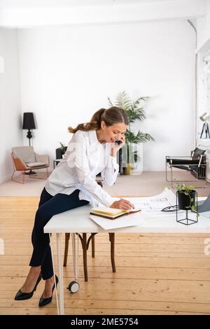Smiling businesswoman talking over mobile phone and making notes sitting on desk in office Stock Photo