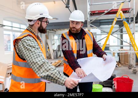 Young engineers wearing protective workwear discussing over blueprint at construction site Stock Photo