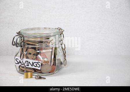 Loose chain on a glass jar with a savings label, money, and open padlock and key on textured light gray background Stock Photo