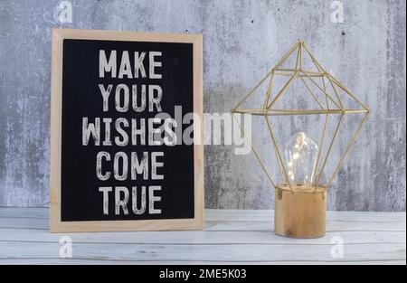 White notebook with inscription MAKE YOUR WISHES COME TRUE written in black pencil on a bright yellow background. Stock Photo