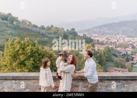 A young family with two children, a teenage girl and a toddler boy, stands against the backdrop of the green hills and vineyards of Italy in the city Stock Photo