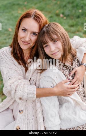 A young caucasian woman with red hair and a teenage daughter are sitting on the green grass, hugging and smiling. They are dressed in knitted clothes Stock Photo