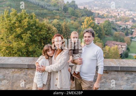 A young family with two children, a teenage girl and a toddler boy, stands against the backdrop of the green hills and vineyards of Italy in the city Stock Photo