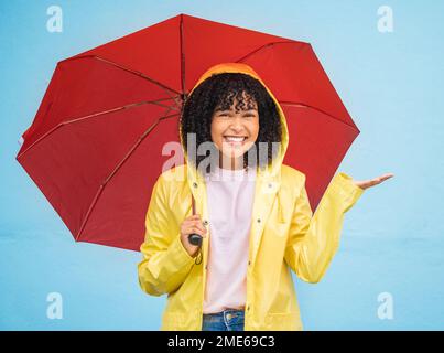 Black woman, portrait and rain umbrella and hand checking for drops on isolated blue background in Brazil city. Happy person, student and raincoat for Stock Photo
