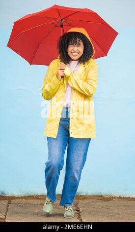 African woman, smile and umbrella by wall with excited face, rain and fashion raincoat in funny moment in city metro. Gen z girl, winter or comic Stock Photo