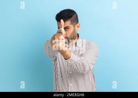 Portrait of dangerous young adult handsome businessman with beard pointing with finger pistols at camera, pretending to shoot, wearing striped shirt. Indoor studio shot isolated on blue background. Stock Photo