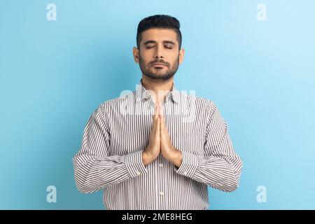 Portrait of calm relaxed young businessman with beard standing doing yoga meditating exercise, keeping palms together, wearing striped shirt. Indoor studio shot isolated on blue background. Stock Photo