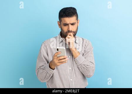 Thoughtful businessman holding chin with smartphone in hand, contemplating about software updating, choosing suitable tariffs, wearing striped shirt. Indoor studio shot isolated on blue background. Stock Photo