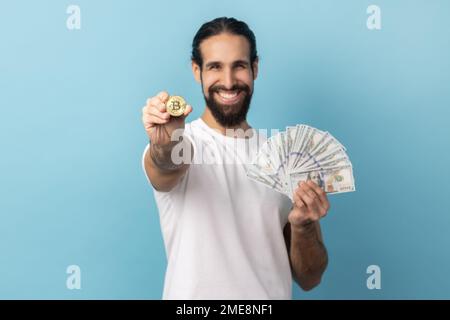 Portrait of happy man with beard wearing white T-shirt showing bitcoin and big fan of dollars banknotes, e-commerce, crypto currency. Indoor studio shot isolated on blue background. Stock Photo