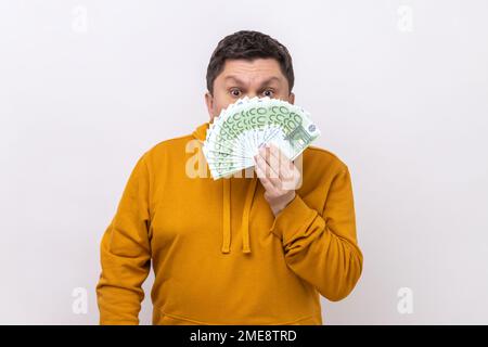 Dark haired man hiding half of face behind fan of hundred euro bills, big winning, betting, wearing urban style hoodie. Indoor studio shot isolated on white background. Stock Photo