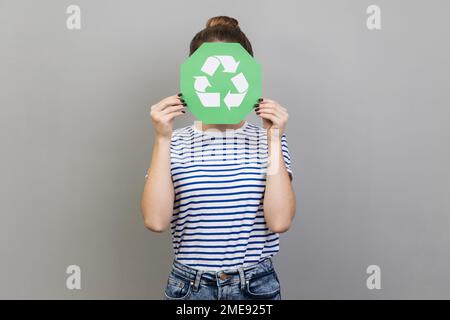 Portrait of anonymous unknown woman wearing striped T-shirt covering her face with green recycling sign, environmental problems. Indoor studio shot isolated on gray background. Stock Photo