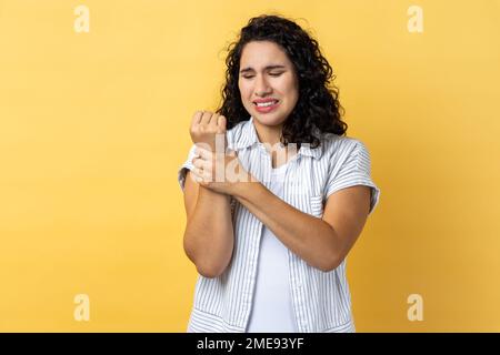 Portrait of unhappy sick woman with dark wavy hair touching painful hand, suffering trauma, sprain wrist, feeling ache of carpal tunnel syndrome. Indoor studio shot isolated on yellow background. Stock Photo
