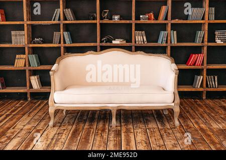 White retro sofa standing in the living room with stylish interior design and collections of books on the bookshelves in the library. Study. Stock Photo
