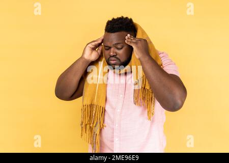 Portrait of unhealthy man wearing pink shirt standing wrapped in scarf, having terrible headache, massaging temples, flu symptoms. Indoor studio shot isolated on yellow background. Stock Photo