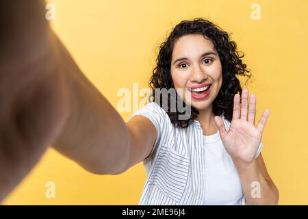 Portrait of smiling friendly woman with dark wavy hair making point of view photo, waving hand, saying hello or good bye, POV. Indoor studio shot isolated on yellow background. Stock Photo