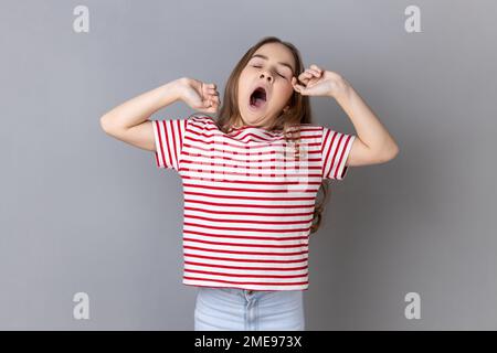 Portrait of sleepy little girl wearing striped T-shirt yawning and raising hands up, feeling fatigued, standing with close eyes. Indoor studio shot isolated on gray background. Stock Photo