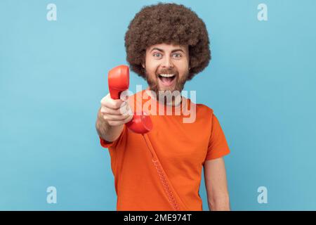 Portrait of man with Afro hairstyle wearing orange T-shirt holding and showing retro phone handset to camera, asking to answer phone. Indoor studio shot isolated on blue background. Stock Photo