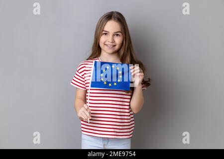 European Union flag. Portrait of smiling adorable cute little girl wearing striped T-shirt holding Europe flag, looking at camera. Indoor studio shot isolated on gray background. Stock Photo