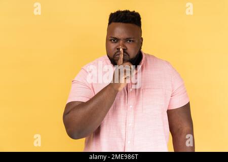 Shh, make silence please. Portrait of serious strict man wearing pink shirt standing showing quiet gesture, saying hush, keep secret. Indoor studio shot isolated on yellow background. Stock Photo
