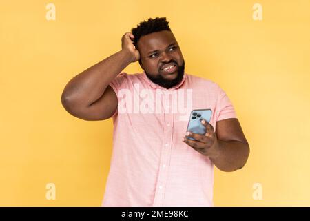 Portrait of pensive thoughtful bearded man wearing pink shirt standing with mobile phone, touching head, having doubt suspicion feeling. Indoor studio shot isolated on yellow background. Stock Photo