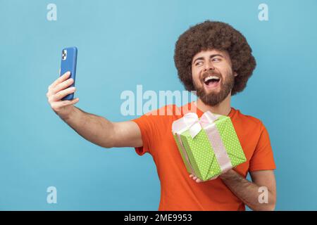 Portrait of happy joyful man with Afro hairstyle wearing orange T-shirt having video call with present box, boasting his gift to followers. Indoor studio shot isolated on blue background. Stock Photo