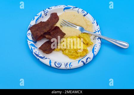 breakfast turkey bacon grits and scrambled eggs on paper plate and light blue background Stock Photo