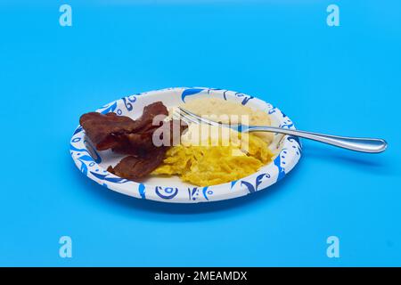 breakfast turkey bacon grits and scrambled eggs on paper plate and light blue background Stock Photo