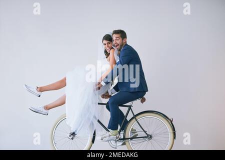 Enjoying lifes ride with my soulmate by my side. Studio portrait of a newly married young couple riding a bicycle together against a gray background. Stock Photo