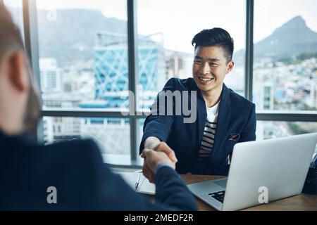 I cant wait to work with you. two young businessmen shaking hands while sitting at a desk in a modern office. Stock Photo