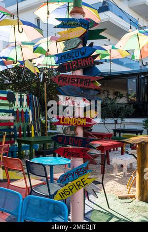 Ksamil, Albania - September 11, 2021: Street wooden sign on the Cafe indicating directions to different places of the world under colorful umbrellas i Stock Photo