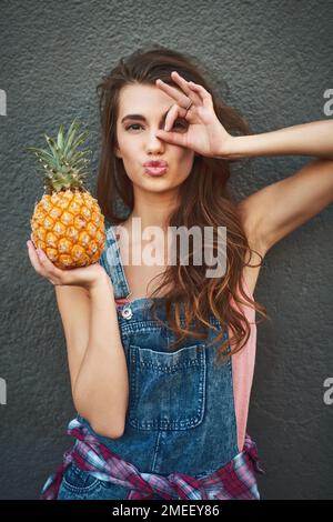 I only see pineapples today. Portrait of a carefree young woman showing a hand gesture on her face while holding a pineapple against a grey background Stock Photo