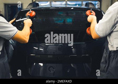 Rear view of two men installing rear window pane into back of car using professional tools. Garage work. Car maintenance. Horizontal indoor shot. High quality photo Stock Photo