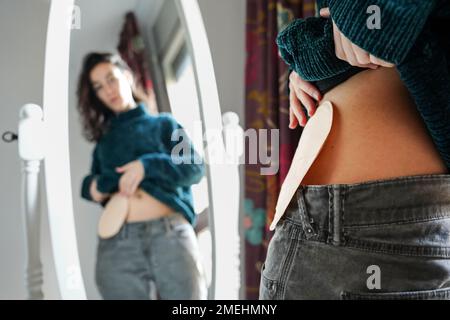 young woman with an ostomy bag looks in the mirror at home. Self-esteem and resilience Stock Photo