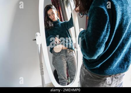 young woman with an ostomy bag on her belly looks in the mirror at home. Self-esteem and resilience after colon cancer Stock Photo