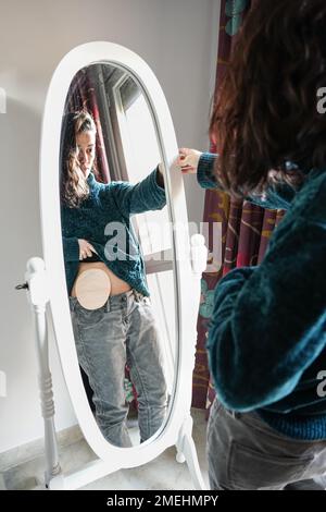 young woman with an ostomy bag on her belly looks in the mirror at home. Self-esteem and resilience after colon cancer Stock Photo