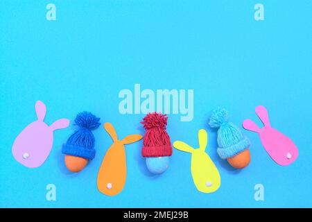 Painted Easter eggs in a winter knitted hat. Rabbit cut out of coloured paper. Blue background. Copy scape. Stock Photo