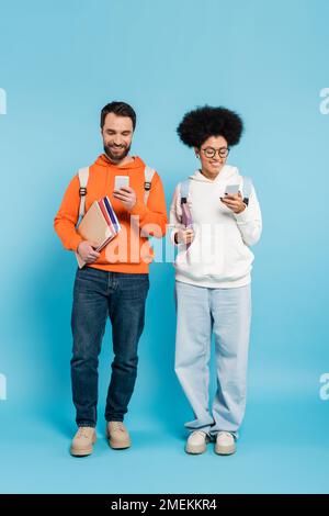 full length of happy multiethnic students with backpacks and copybooks using smartphones on blue Stock Photo